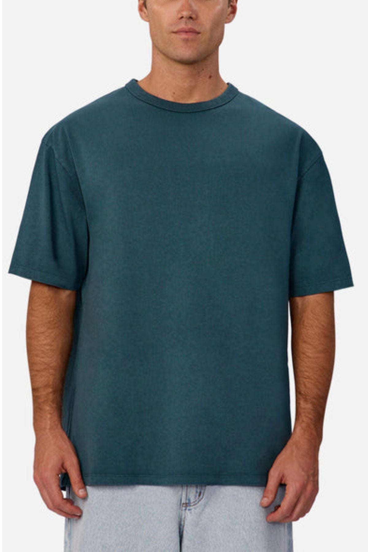 A man stands against a plain background facing forward, wearing an oversized Industrie The Del Sur Tee in teal and light blue jeans, with his arms relaxed at his sides.