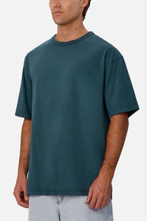 A man wearing the Industrie The Del Sur Tee, a relaxed heavyweight cotton t-shirt in teal, and light blue jeans stands against a plain white background. His face is partially out of frame with his arms at his sides.