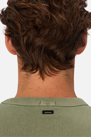 The image shows the back of a person with wavy brown hair wearing an oversized sage green Industrie The Del Sur Tee made of heavyweight fabric, featuring a small black "Industrie" label on the collar.