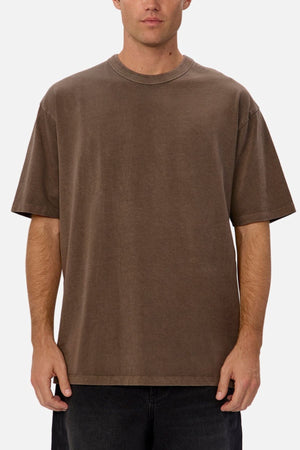 An individual stands facing forward against a plain background, wearing the Industrie The Del Sur Tee in brown—a loose-fitting, heavyweight cotton t-shirt with a simple crew neckline—paired with dark pants and featuring no visible logos or designs.