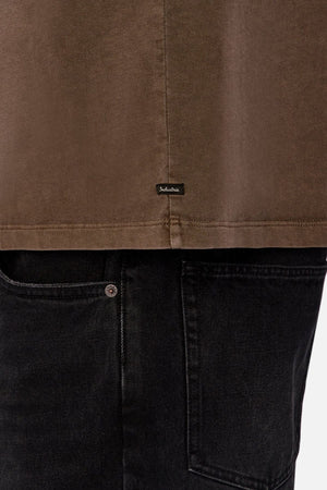 A close-up of a person wearing the Industrie The Del Sur Tee in brown—a heavyweight, oversized cotton tee—paired with black jeans, showcasing the shirt's small black label and the upper part of the jeans pocket and waistband.