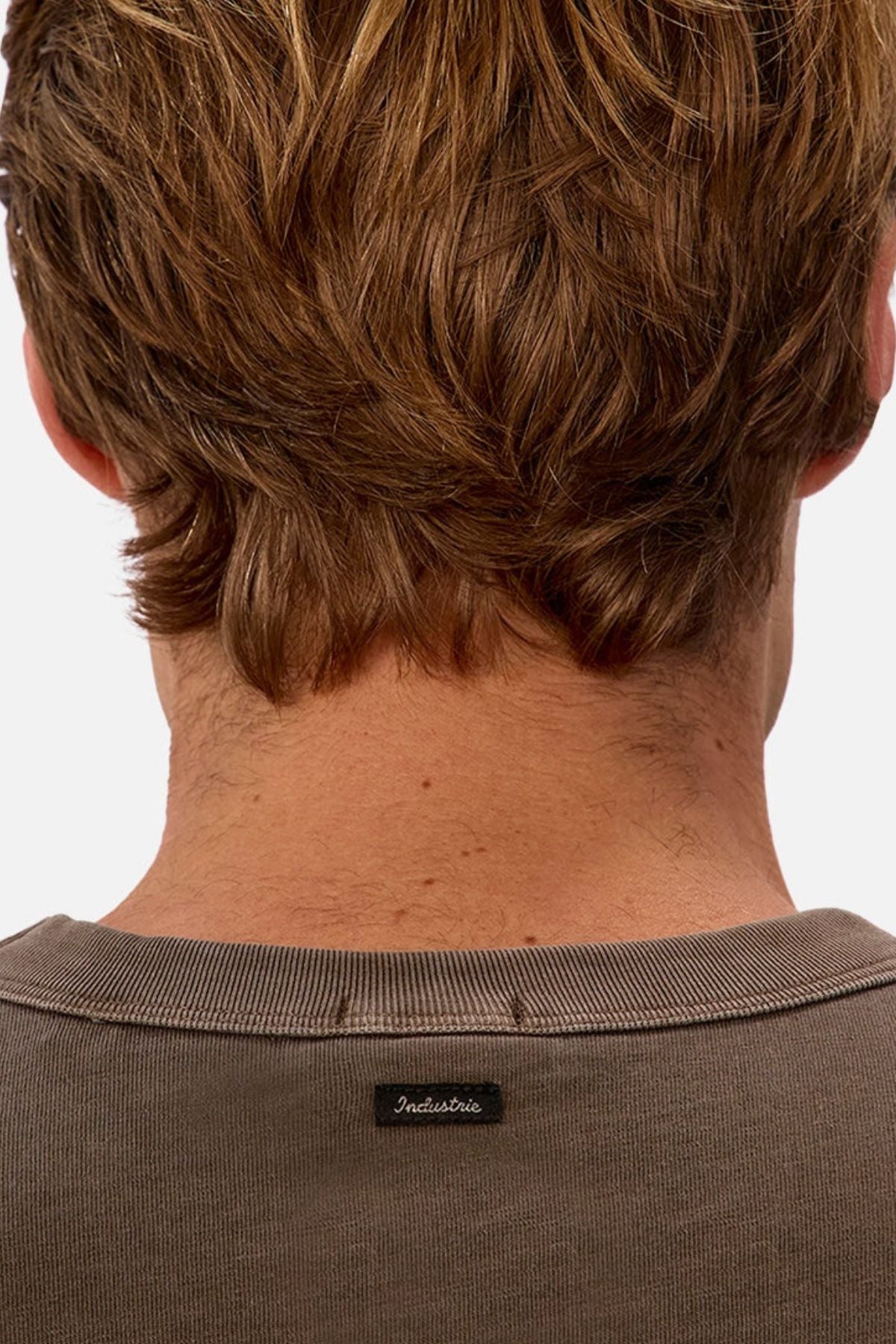 The image displays the back of a person with light brown, wavy hair wearing an Industrie The Del Sur Tee—a heavyweight cotton tee with a small black collar label—against a plain, light background.