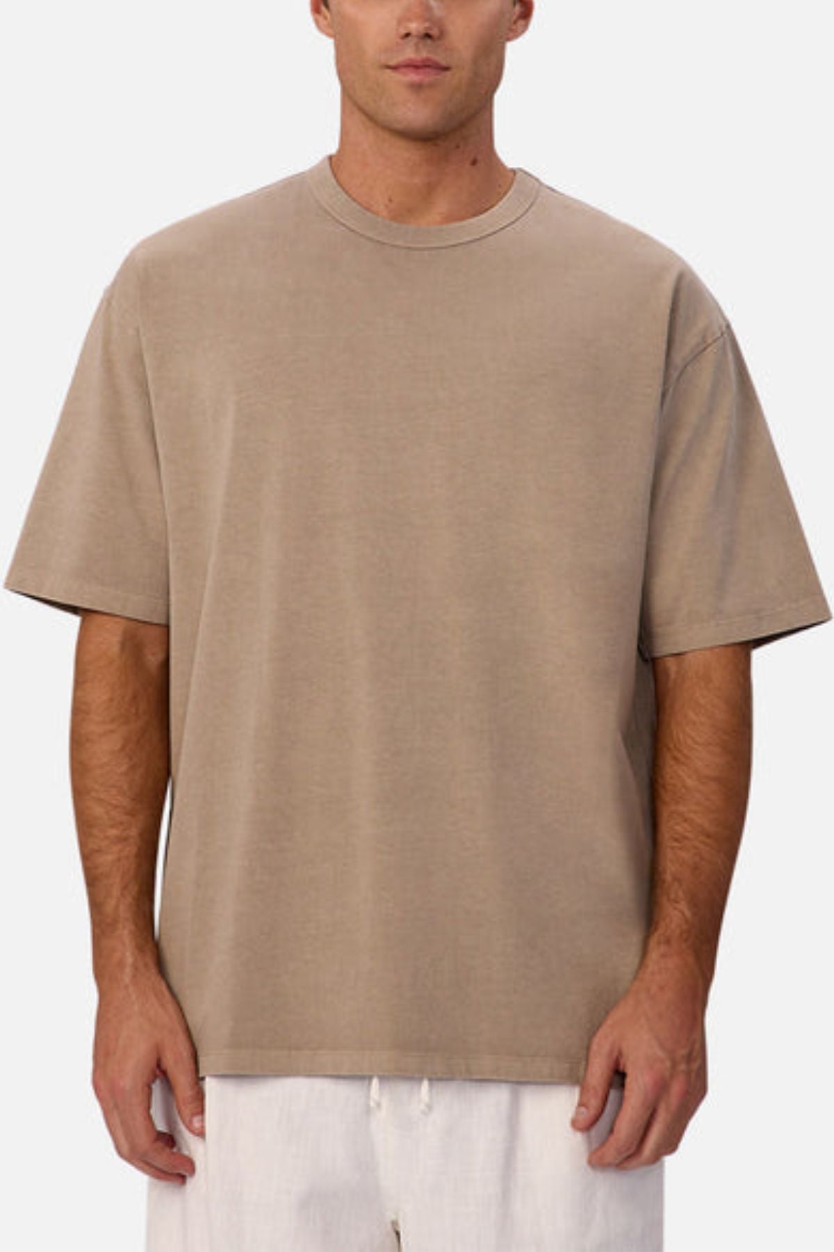 A man wears an oversized Industrie The Del Sur Tee in beige with white pants, standing against a plain white background. His face is partially visible.
