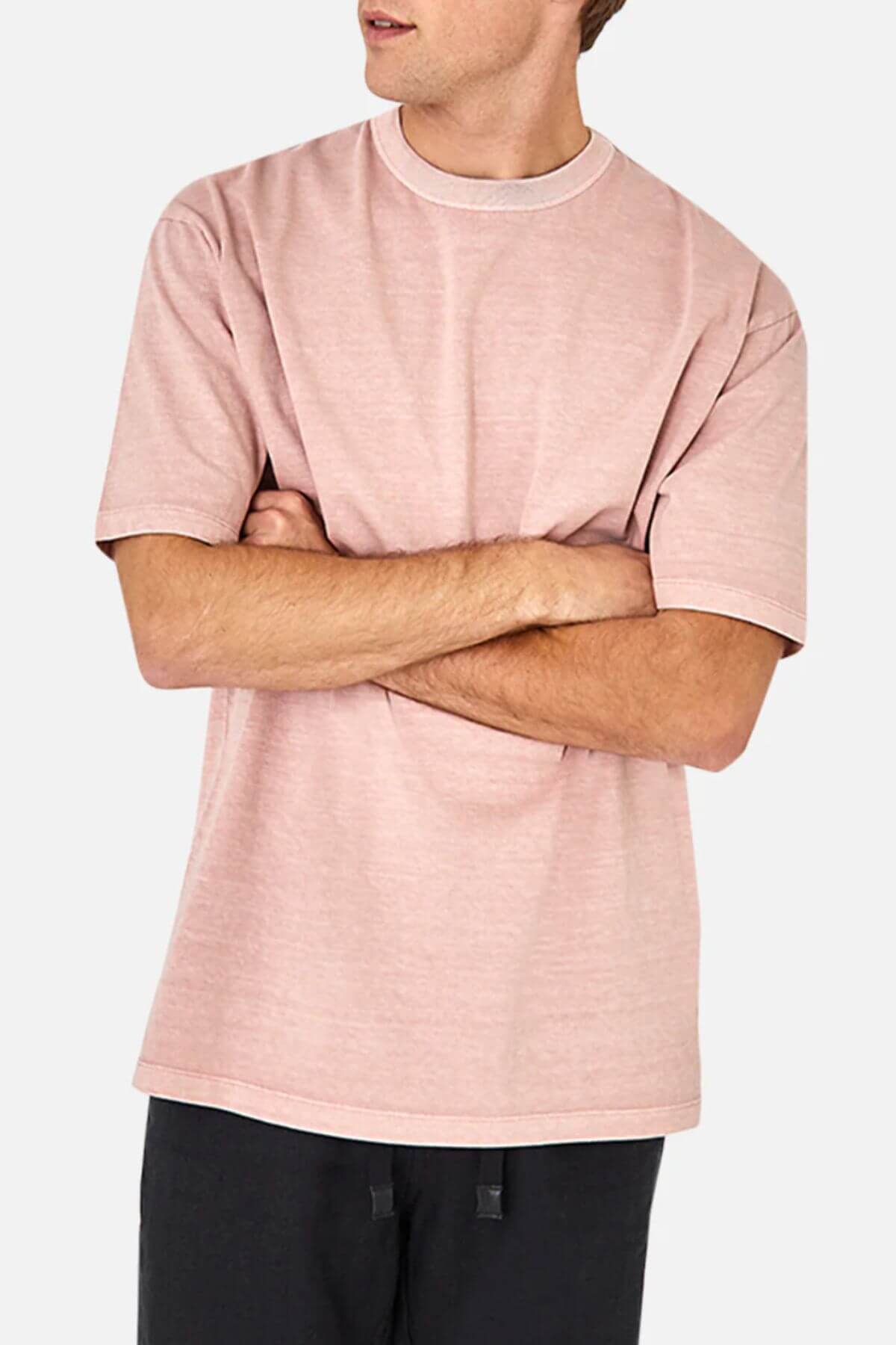 Person in an Industrie The Del Sur Tee, light pink and comfy with an oversized fit and short sleeves, paired with black pants, stands against a plain background, arms crossed and facing slightly left.