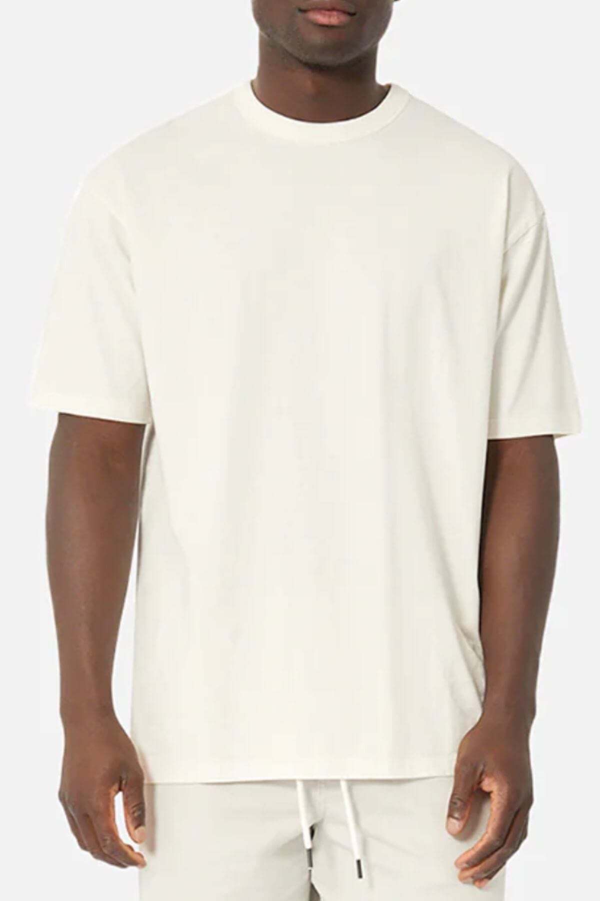 A person wearing an oversized Industrie The Del Sur Tee, made from 100% cotton, stands against a gray background. The off-white T-shirt features short sleeves and a loose fit, paired with light-colored pants, highlighting the simplicity of the outfit.