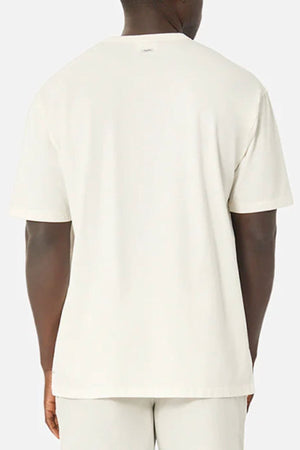 A person stands against a neutral background wearing an Industrie The Del Sur Tee, a plain, light-colored oversized T-shirt made from 100% cotton. It pairs with matching pants. The shirt is loose-fitting, and the fabric looks incredibly soft.