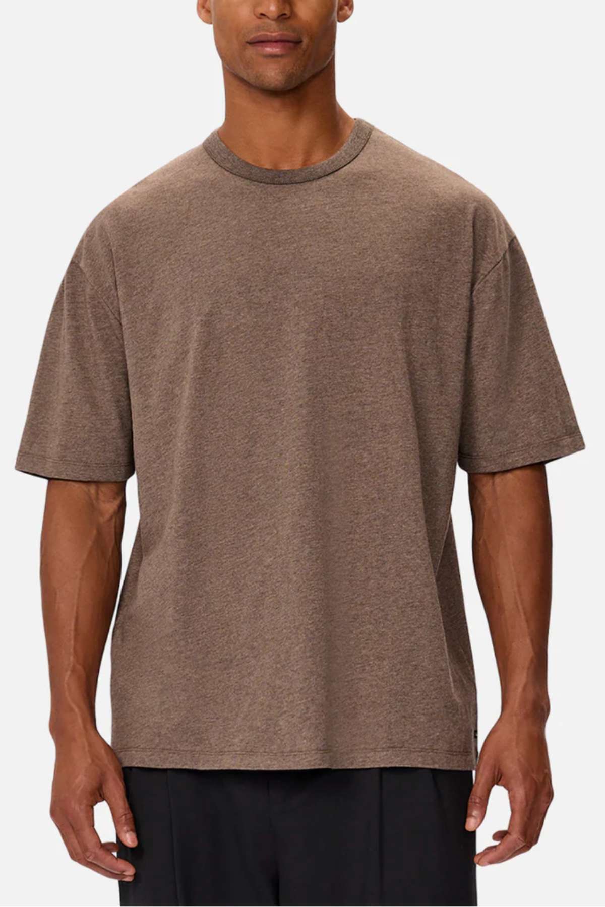 A person in an oversized Industrie The Del Sur Tee, made of heavyweight fabric and featuring a simple crew neckline, stands with relaxed arms against a plain backdrop. The brown T-shirt pairs effortlessly with black pants for a stylish look.