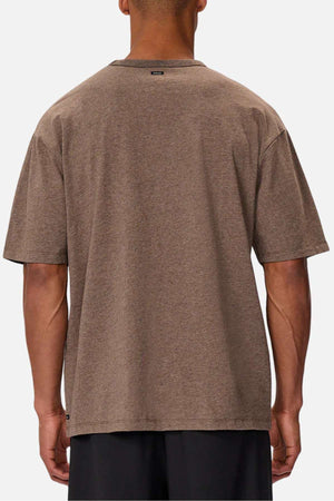 A person in an Industrie The Del Sur Tee, plain brown, stands with their back to the camera against a white background. The heavyweight fabric offers sturdiness, and a black tag near the neckline adds a subtle detail.