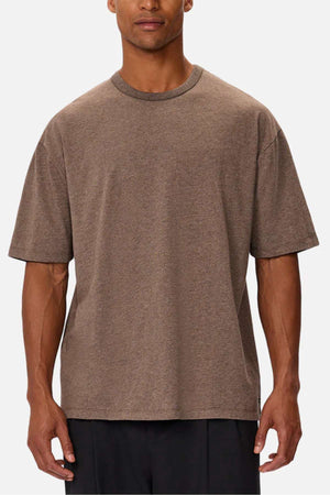 A person in an oversized Industrie The Del Sur Tee, made of heavyweight fabric and featuring a simple crew neckline, stands with relaxed arms against a plain backdrop. The brown T-shirt pairs effortlessly with black pants for a stylish look.