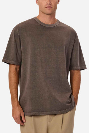 A person wearing an oversized fit, faded brown "Industrie The Del Sur Tee" paired with beige pants is standing against a plain background. The T-shirt, crafted from 100% cotton, boasts a washed, vintage appearance, while the person casually rests one hand in their pocket.