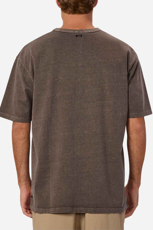 The person is showcasing an oversized fit, 100% cotton brown T-shirt—Industrie The Del Sur Tee—with a subtle tag at the neck on the back. Complemented by light-colored pants, they stand facing away from the camera, embodying a relaxed and stylish vibe.