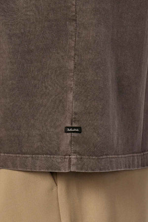 Close-up of the hem of The Del Sur Tee by Industrie, a brown oversized fit shirt made from 100% cotton, with a small black tag displaying a white logo. The tee is styled with beige pants.