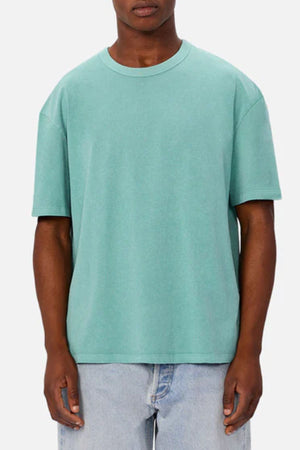 A person in the Industrie The Del Sur Tee, a loose-fitting short-sleeve light teal oversized t-shirt, paired with light blue jeans stands facing forward against a plain gray background. Their face is partially obscured.