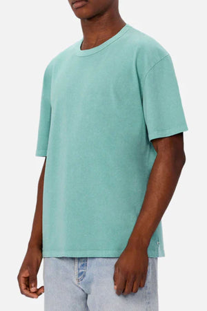 A person in an oversized Industrie The Del Sur Tee in plain teal and light blue jeans stands before a light grey backdrop. The relaxed fit and partially out-of-frame head create a modern, minimal style.