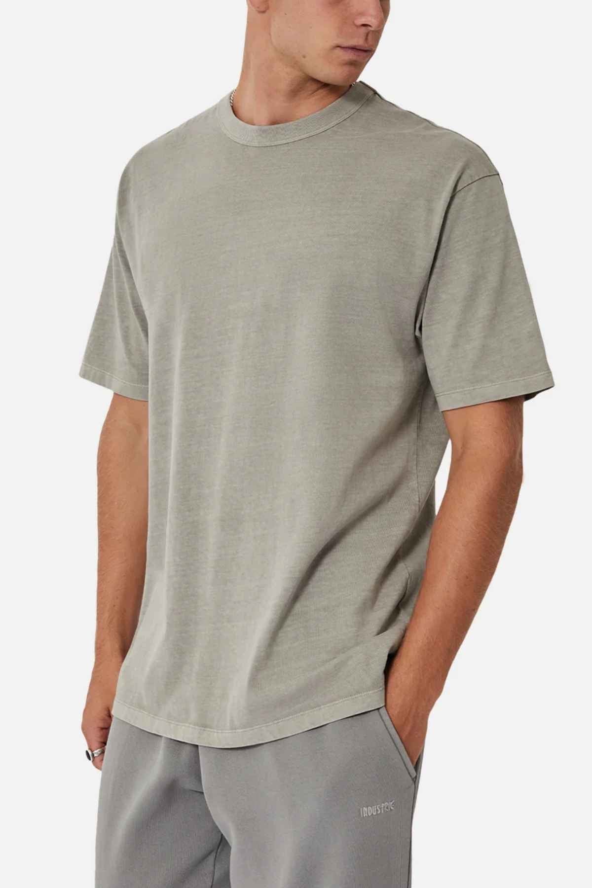 The man is wearing the Industrie The Del Sur Tee in light gray, made of 100% cotton, paired with pocketed sweatpants. Standing against a plain white background, he looks to his right with a casually cool vibe, his left hand resting in his pocket.
