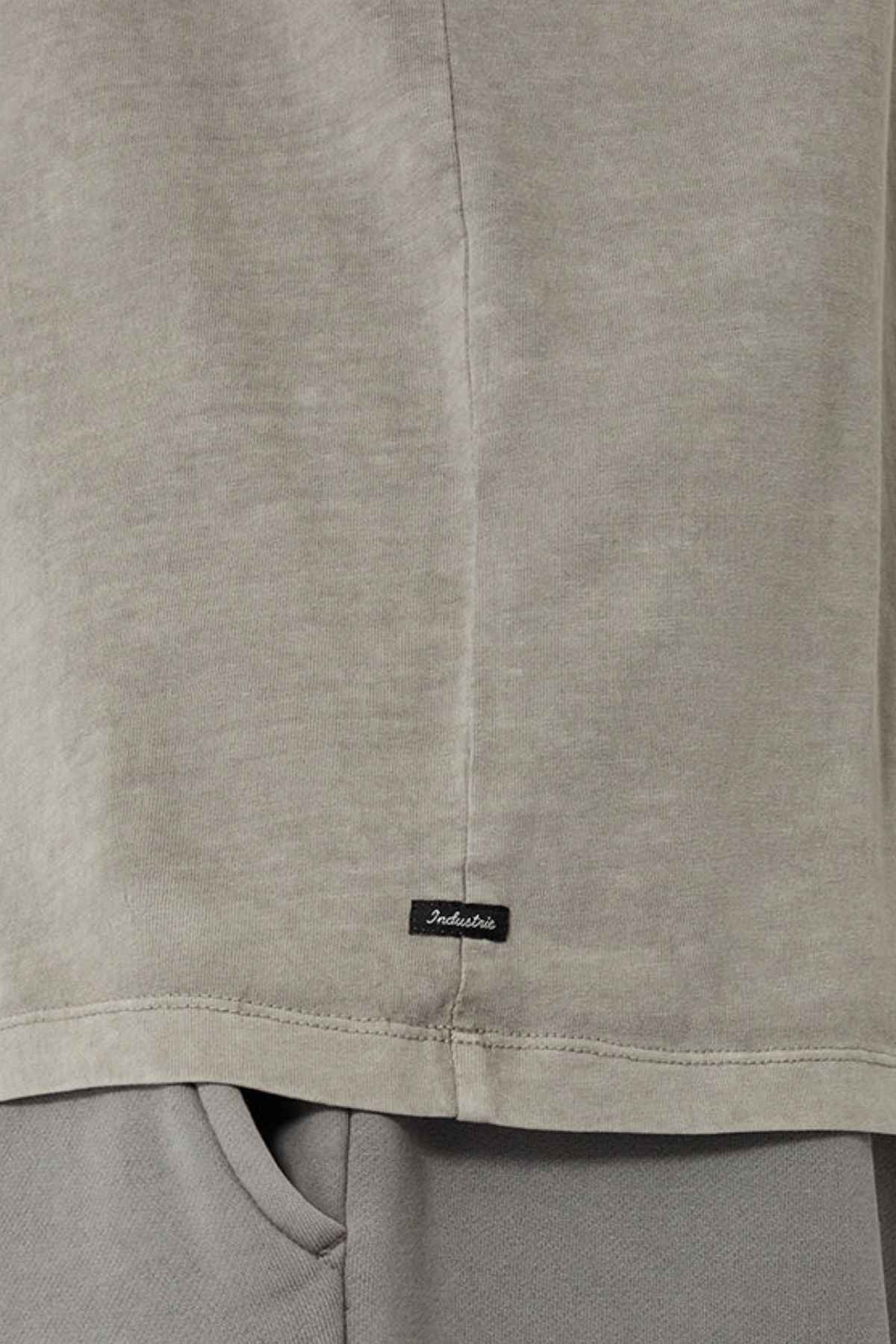 A close-up view of a light gray 100% cotton fabric highlights the hem of the oversized Industrie The Del Sur Tee, adorned with a small black tag featuring indistinct text. The tee elegantly overlays gray pants, achieving a refined monochromatic aesthetic.