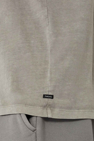 A close-up view of a light gray 100% cotton fabric highlights the hem of the oversized Industrie The Del Sur Tee, adorned with a small black tag featuring indistinct text. The tee elegantly overlays gray pants, achieving a refined monochromatic aesthetic.