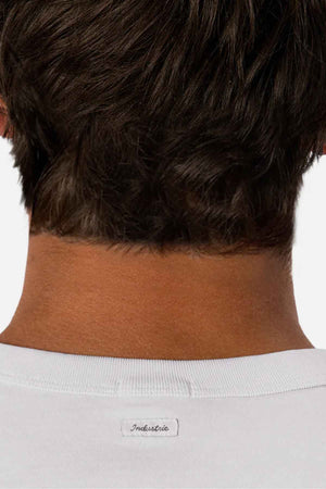 The image features the back of a person's head and neck with short brown hair. They are wearing a white, comfortable tee called "Industrie The Del Sur Tee," made from 100% cotton, with a small label displaying the word "Industrie.