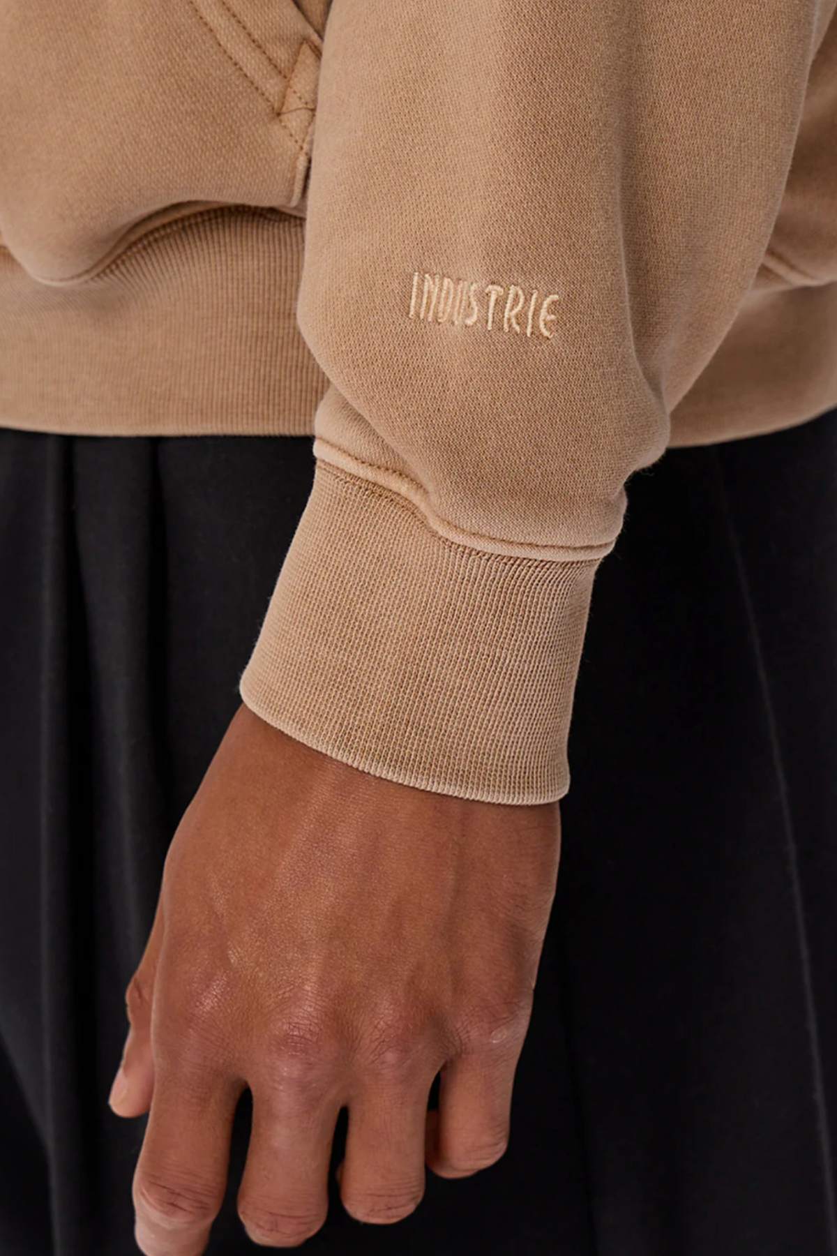 Close-up of a person wearing the Industrie The Del Sur Washed Hoodie in an oversized camel fit, featuring "INDUSTRIE" embroidered on the sleeve, paired with black pants.
