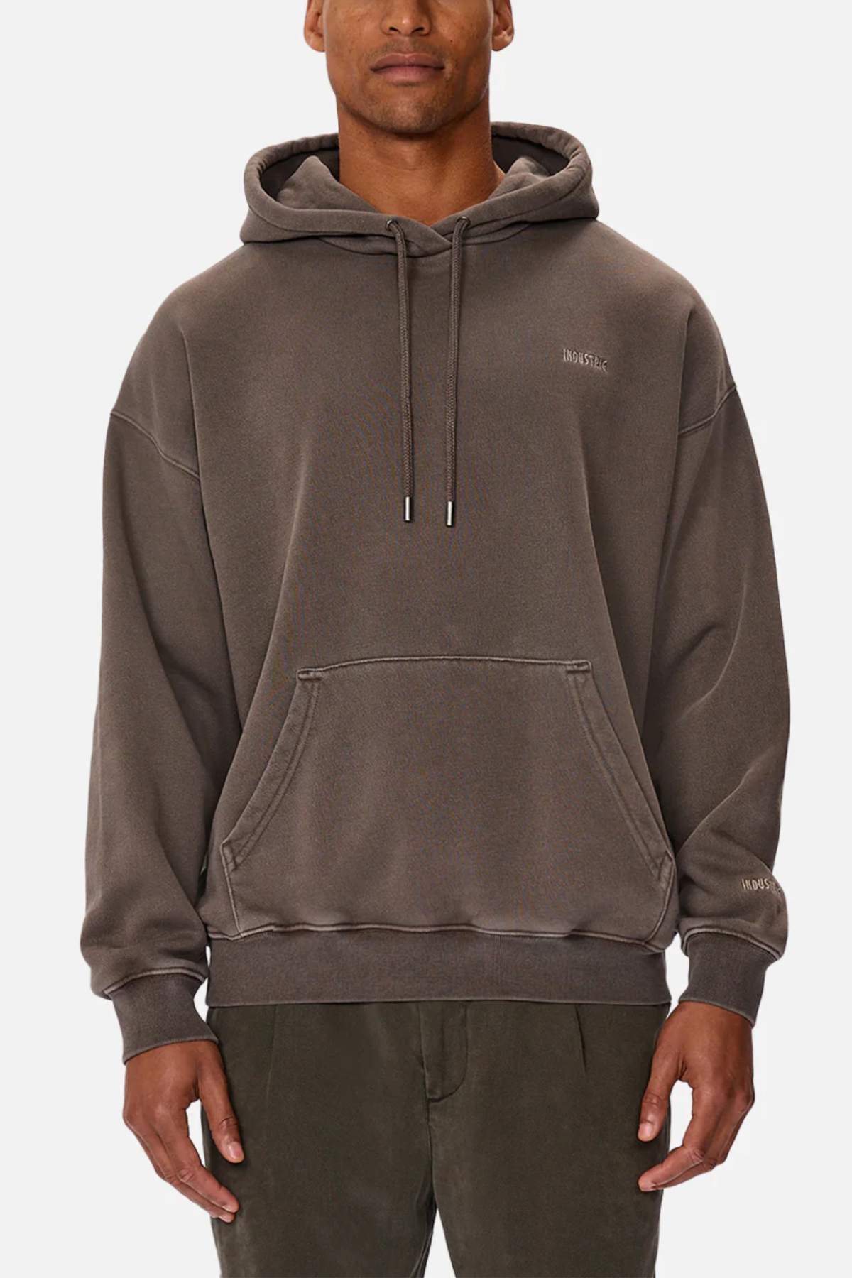 A person in a loose-fitting, acid wash hoodie and dark pants stands against a white background. The Industrie "The Del Sur Washed Hoodie" features a front pocket, drawstrings, and subtle embroidered text on the chest and sleeve.