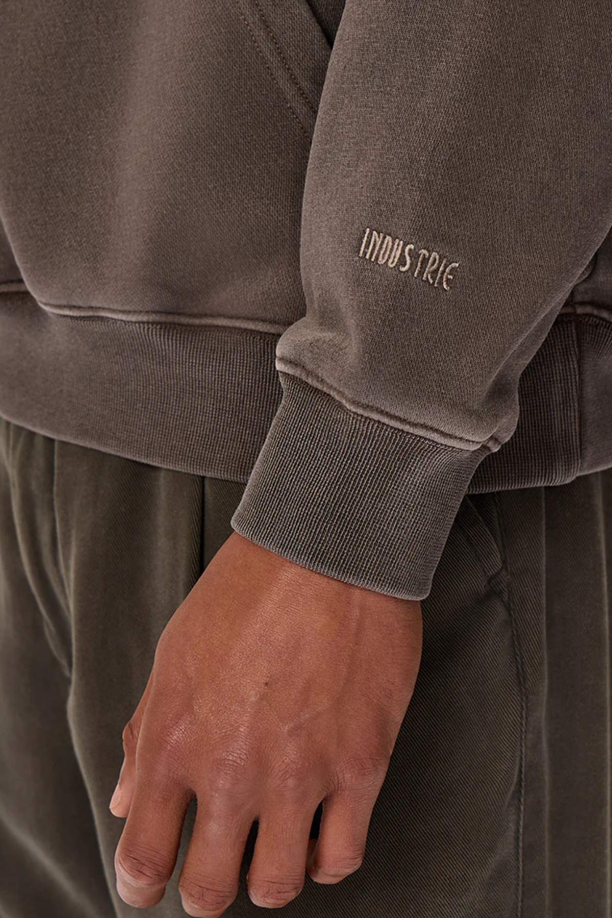 Close-up of a person's hand by their side, wearing the Industrie The Del Sur Washed Hoodie, an acid wash brown sweatshirt with "INDUSTRIE" on the sleeve. Fabric texture and seam details are visible. Matching pants complete the outfit.