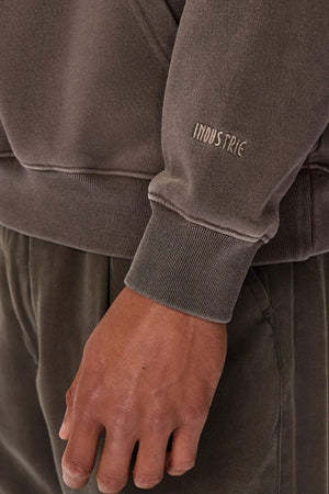 Close-up of a person's hand by their side, wearing the Industrie The Del Sur Washed Hoodie, an acid wash brown sweatshirt with "INDUSTRIE" on the sleeve. Fabric texture and seam details are visible. Matching pants complete the outfit.