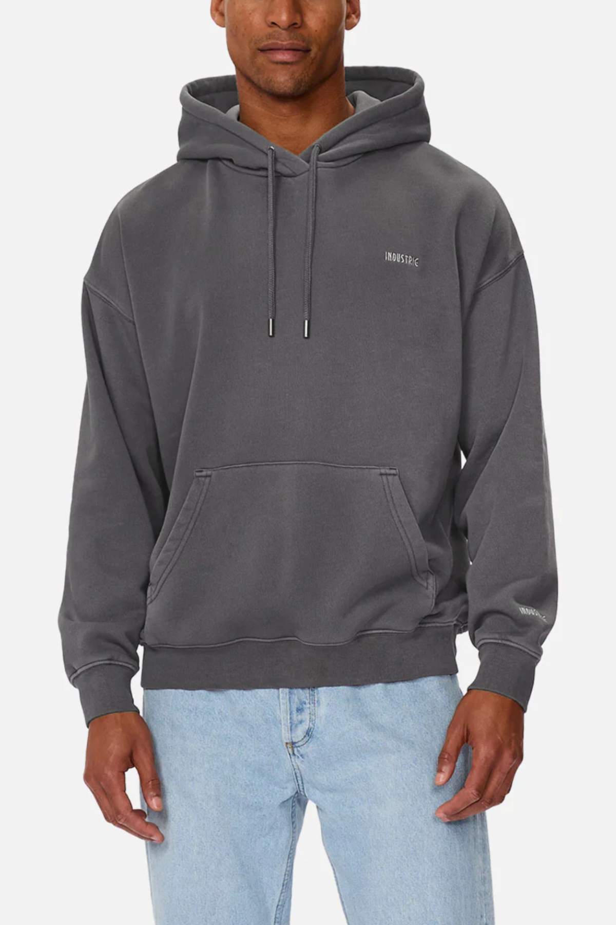 A person wears the Industrie The Del Sur Washed Hoodie in an oversized fit, dark gray color with a front pocket, paired with light blue acid wash jeans, standing against a plain white background.