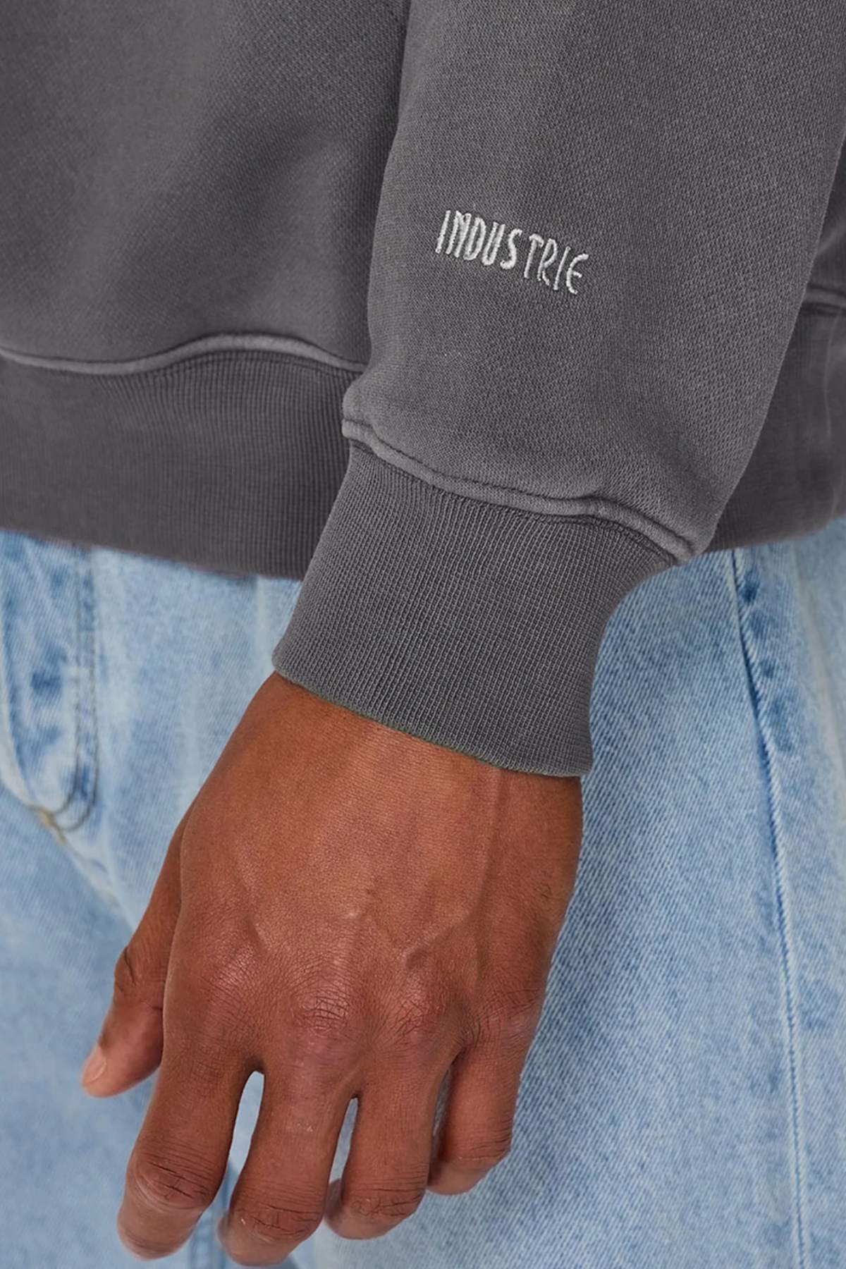 A close-up of a person wearing the Industrie The Del Sur Washed Hoodie in dark gray, featuring "INDUSTRIE" embroidery on the sleeve, paired with light blue denim jeans. Their hand rests casually near the seam of the jeans.