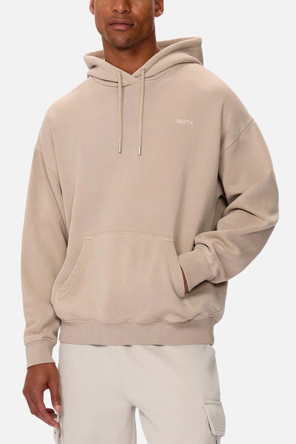 A man wears the oversized Industrie The Del Sur Washed Hoodie, subtly printed with "DISTRICT" on the chest. It features a front pocket and drawstrings, perfectly complementing his light-colored pants as he stands against a plain background.