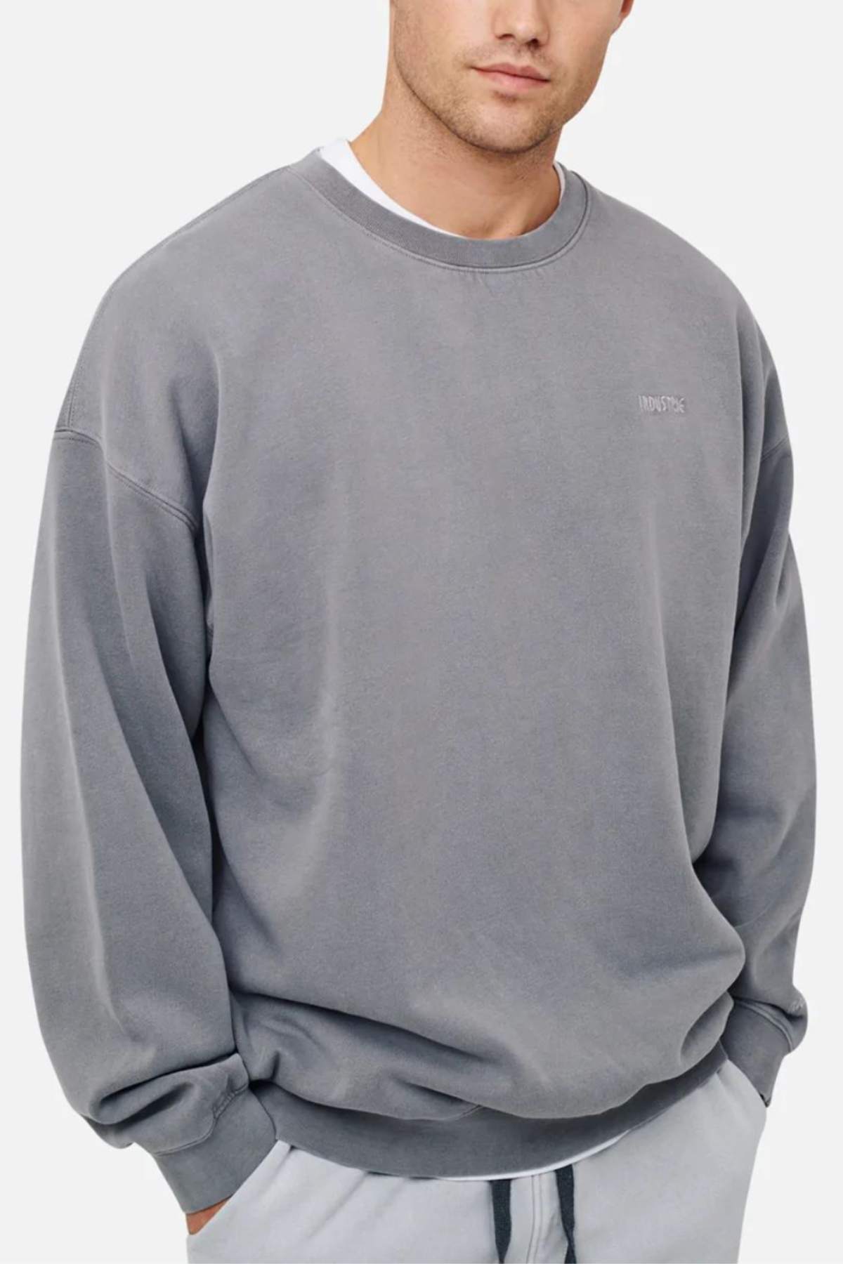 Dressed in the Industrie The Del Sur Washed Sweat, a person layers this oversized gray jumper with chest text over a white shirt, paired with light gray pants. Set against a plain white backdrop, the ensemble exudes a cozy and casual vibe.