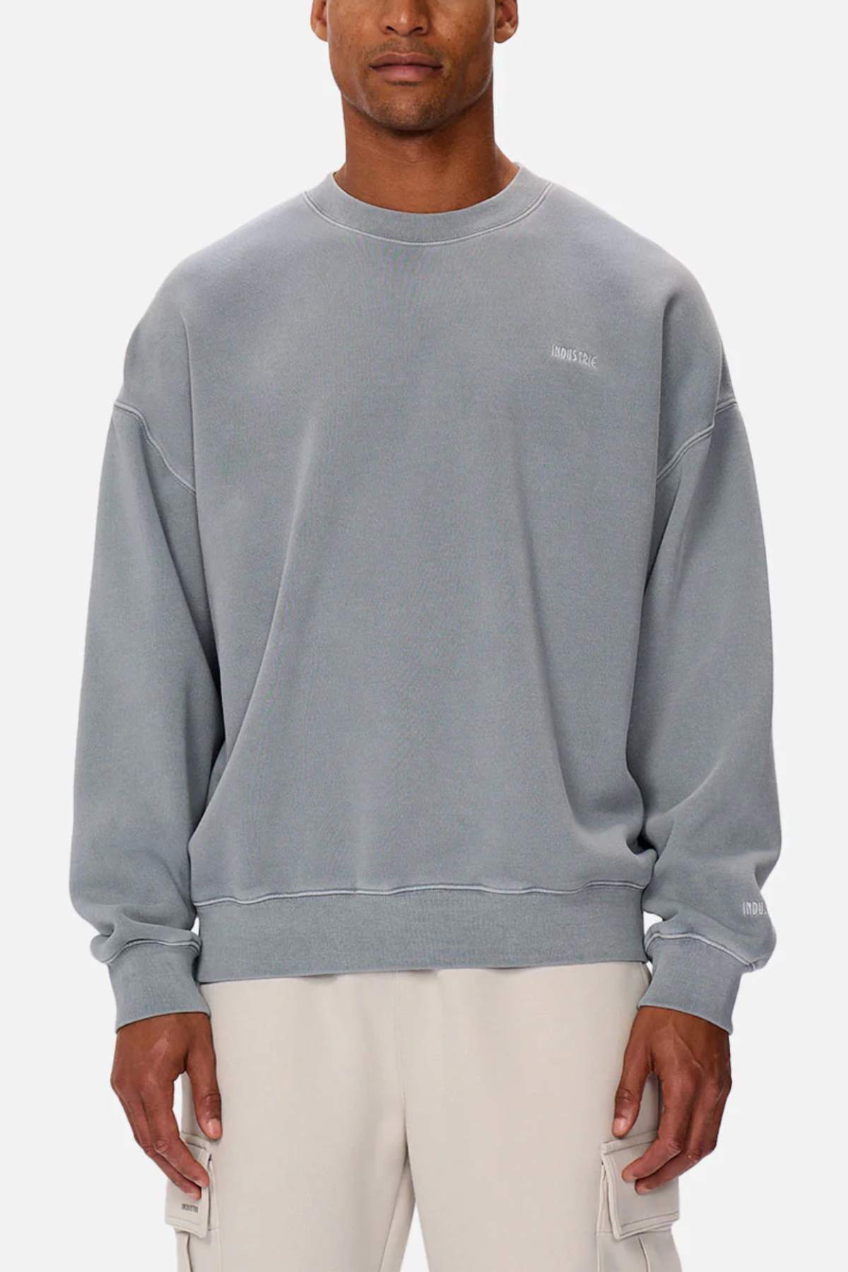 A man wears the Industrie The Del Sur Washed Sweat, an oversized faded gray jumper with subtle white chest and sleeve text and rib knit cuffs, paired with light cargo pants, standing against a plain background.