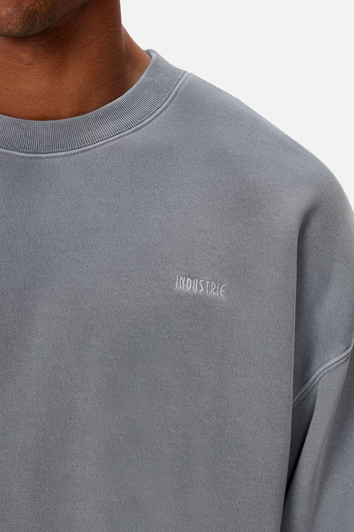 A close-up of a person wearing the Industrie The Del Sur Washed Sweat in grey, featuring "INDUSTRIE" embroidered in small white letters on the left chest and rib knit cuffs, with only the lower face, neck, and upper torso visible.