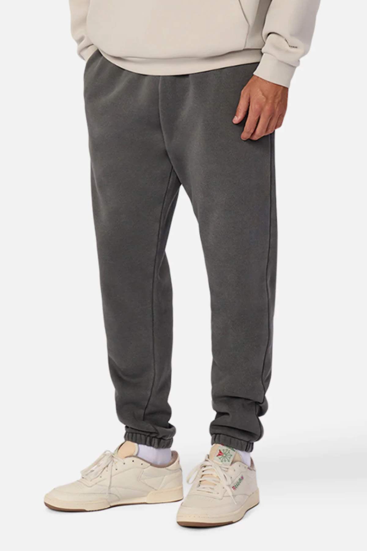 A person wears Industrie The Del Sur Washed Track Pant in charcoal gray with an elasticated drawstring waist, a beige sweatshirt, and off-white sneakers with pastel accents, standing against a plain white background.