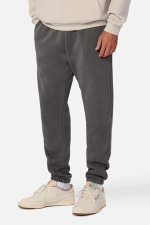 A person wears Industrie The Del Sur Washed Track Pant in charcoal gray with an elasticated drawstring waist, a beige sweatshirt, and off-white sneakers with pastel accents, standing against a plain white background.