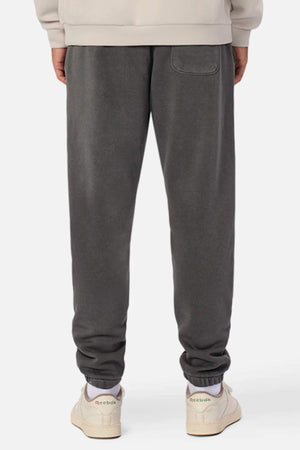 Rear view of a person wearing dark gray Industrie The Del Sur Washed Track Pant with an elastic drawstring waist and single back pocket, paired with a beige sweatshirt and off-white Reebok sneakers, standing against a plain white background.