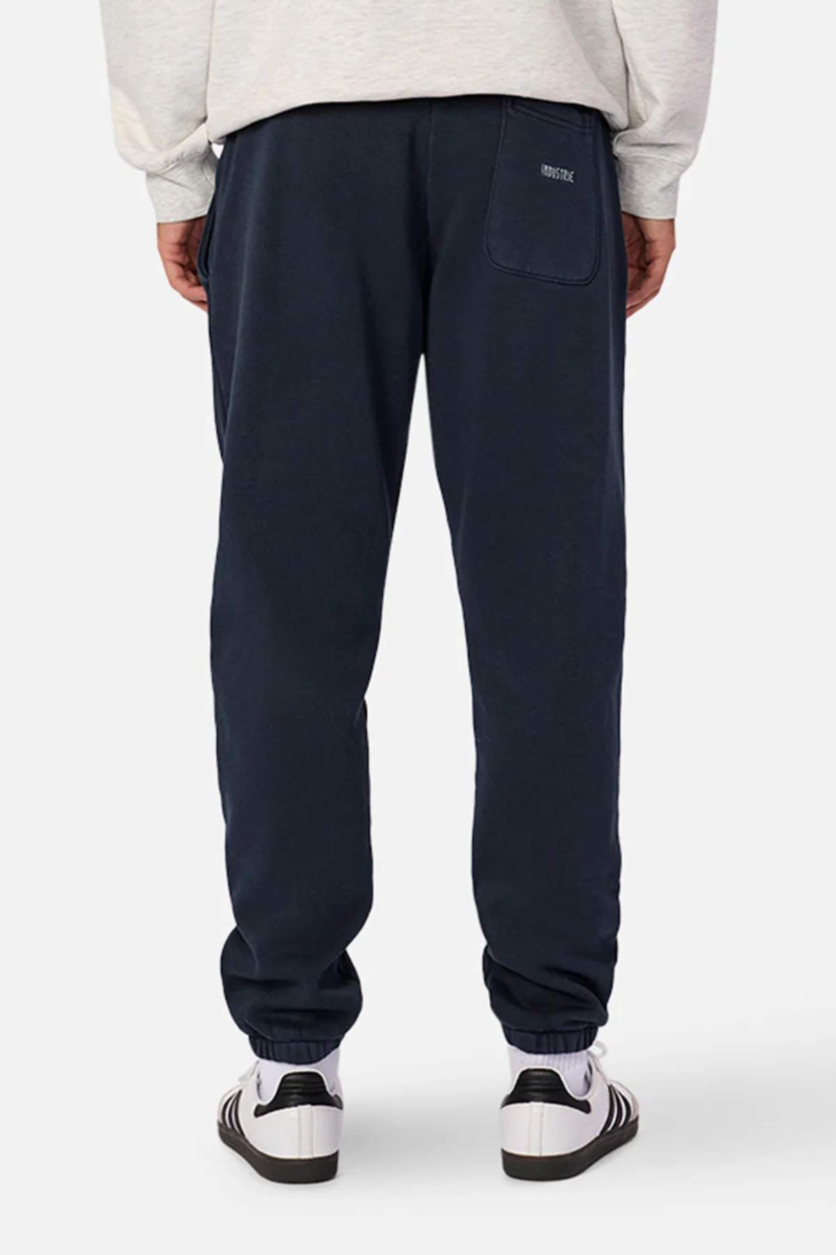 Back view of a person wearing the Industrie The Del Sur Washed Track Pant in navy blue, featuring an elasticated drawstring waist and a back pocket, paired with white sneakers and an oversized fit light-colored sweatshirt. The jogger pants have elastic cuffs and the brand "DISTRICT" printed above the pocket. The background is plain white.