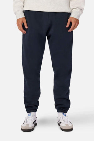 A person is standing against a plain background, wearing an oversized light-colored sweatshirt and the Industrie The Del Sur Washed Track Pant, which are dark blue sweatpants featuring an elasticated drawstring waist. The person is also wearing white athletic shoes with blue accents. The image showcases the lower half of the person's body, from the waist down to their feet.