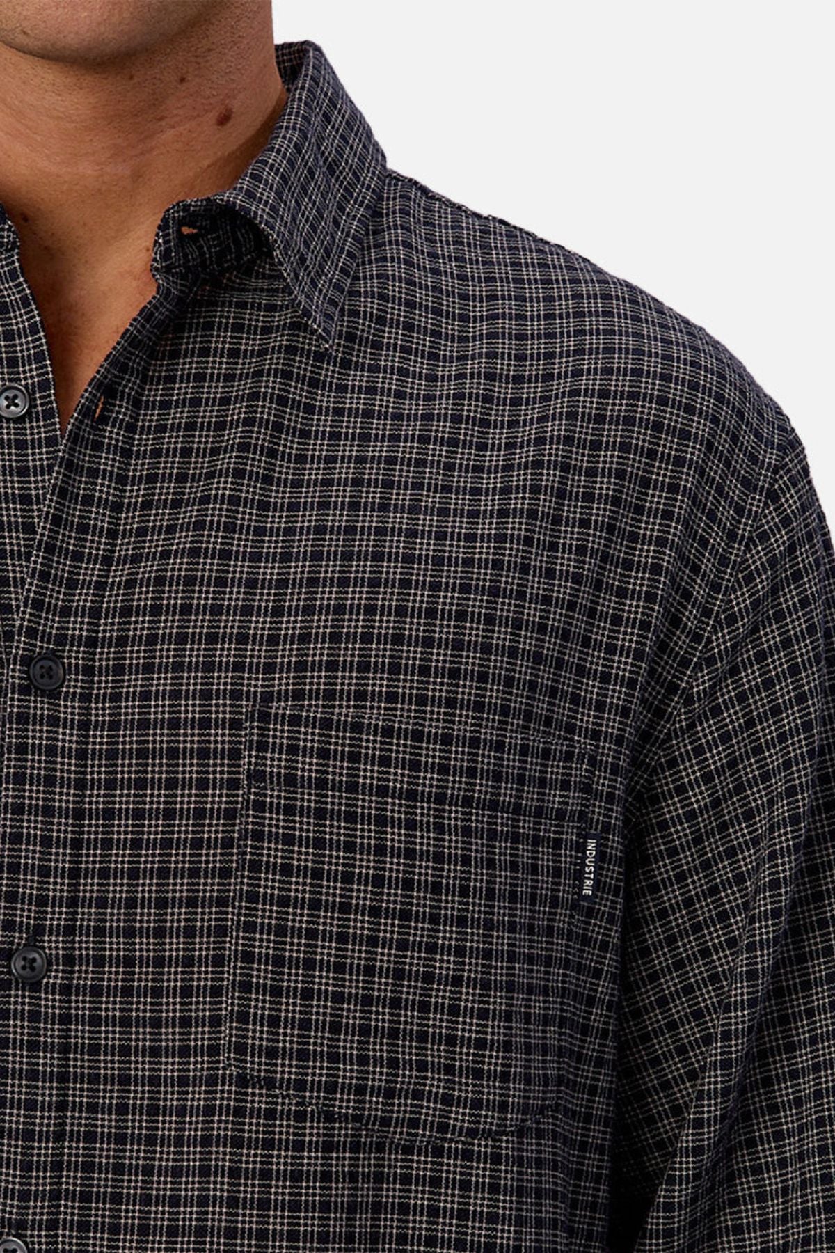 A close-up of a person wearing the Industrie The Diego Long Sleeve Shirt, featuring a dark checked pattern and front pocket. Only the lower face, neck, shoulder, and upper chest are seen against a white background.