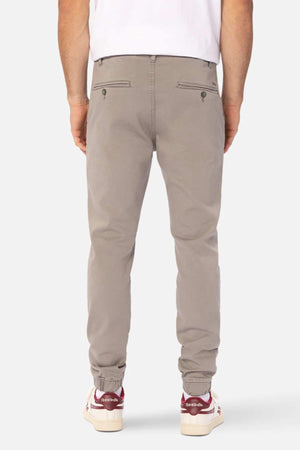 Someone is shown from the back wearing Industrie The Drifter Chino Pant in a slim fit light gray style with an elastic waist, paired with a white T-shirt and red-and-white Reebok sneakers.