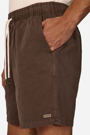 A person is shown wearing the Industrie The Dublin Short, a summer staple with an elasticised waistband and a white drawstring. One hand is tucked into the pocket, while a small brand label is visible near the hem against a plain white background.