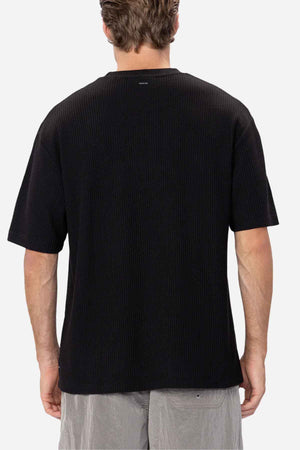 A person with short hair is standing facing away from the camera, wearing the Industrie The Earl Waffle Tee in black, which features a loose fit and a classic crew neck, paired with light gray pants. The background is plain white.