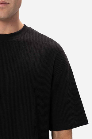 A close-up of a person wearing the Industrie The Earl Waffle Tee, a black, textured short-sleeve shirt featuring a ribbed pattern and a classic crew neck. Only the upper torso and part of the face are visible, set against a plain light background.