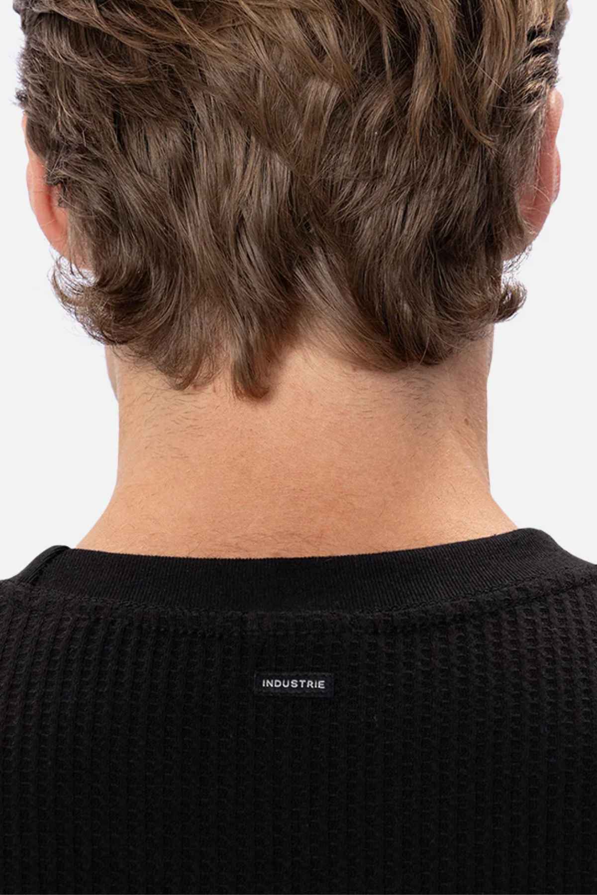 Close-up of the back of a person's head with wavy brown hair, wearing the Industrie The Earl Waffle Tee in black. The word "INDUSTRIE" is written in small white letters below the neckline. The background is plain white.