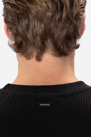 Close-up of the back of a person's head with wavy brown hair, wearing the Industrie The Earl Waffle Tee in black. The word "INDUSTRIE" is written in small white letters below the neckline. The background is plain white.