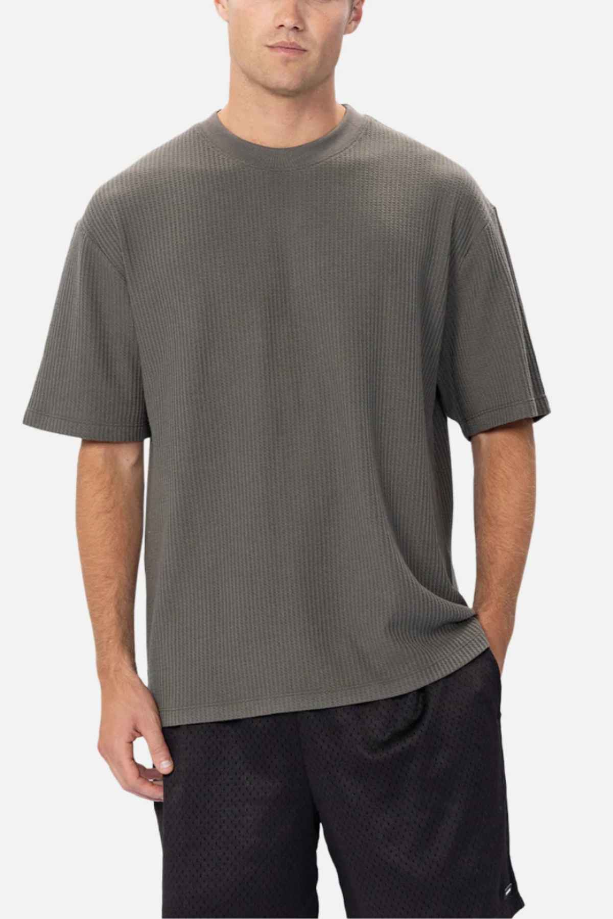 A person stands against a plain background wearing an oversized, dark green Industrie The Earl Waffle Tee and black shorts. The individual's hands are in their pockets, and they have a neutral expression.