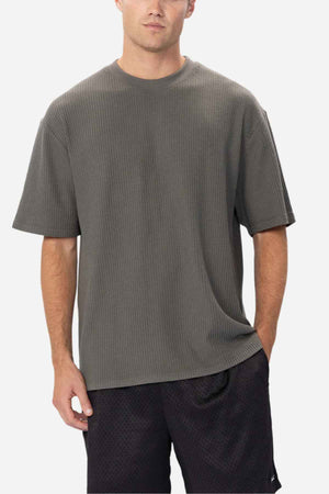 A person stands against a plain background wearing an oversized, dark green Industrie The Earl Waffle Tee and black shorts. The individual's hands are in their pockets, and they have a neutral expression.