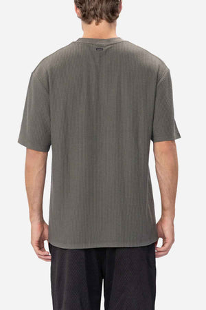 A person with short hair is standing with their back to the camera, wearing a loose-fitting, dark grey Industrie The Earl Waffle Tee and black athletic shorts. The classic crew neck design complements the comfortable fabric blend. The background is plain white.