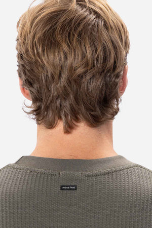 Back view of a person with wavy, light brown hair wearing a long-sleeved, green ribbed Industrie The Earl Waffle Tee. The focus is on the hair and the shirt's texture, with a small rectangular tag on the neckline reading "INDUSTRIE." The background is plain white.