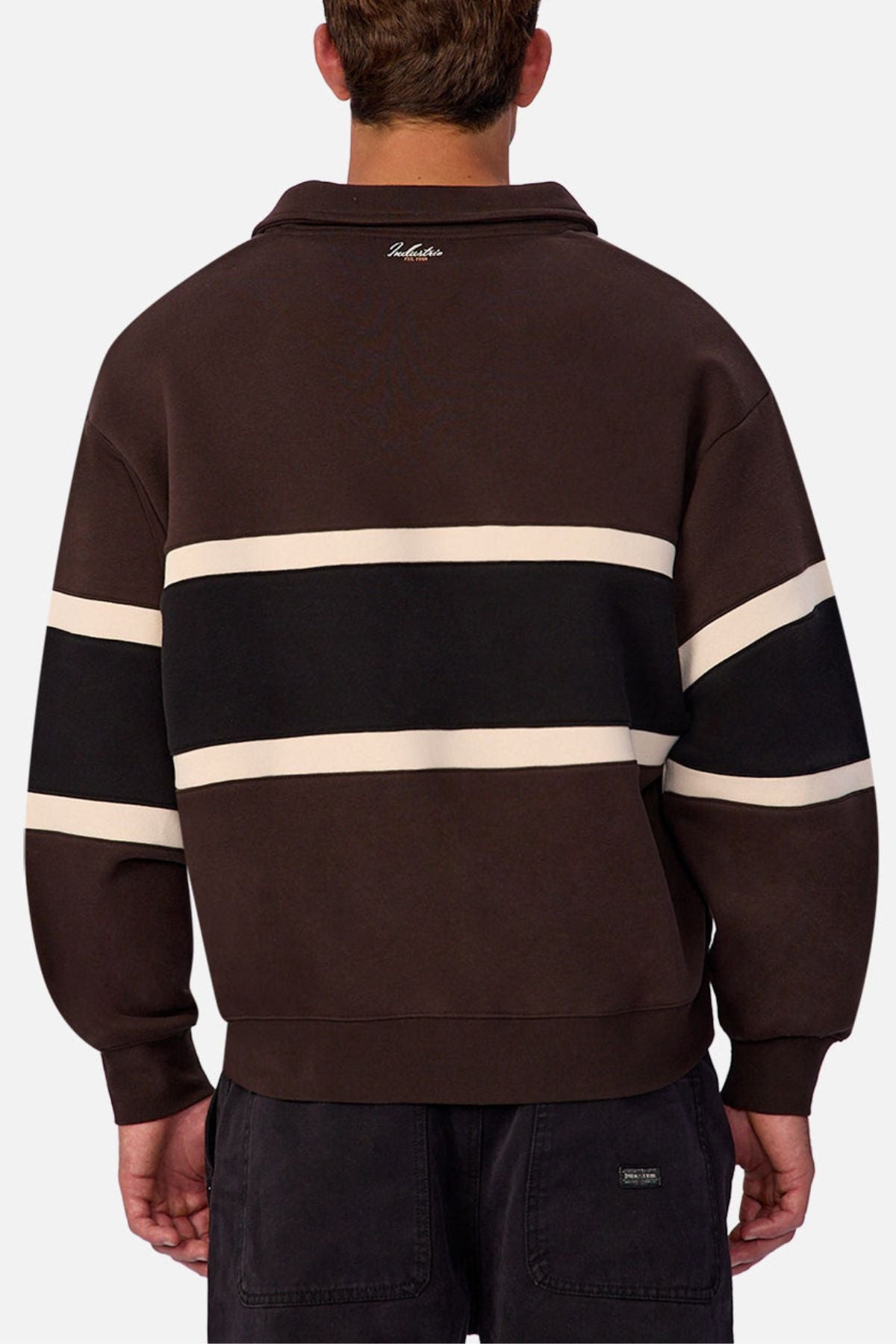 Someone is wearing the Industrie The Eastbourne Tracktop in dark brown, featuring black and white horizontal stripes on the back, paired with dark pants. The photo captures the relaxed silhouetteโs back view against a light background.
