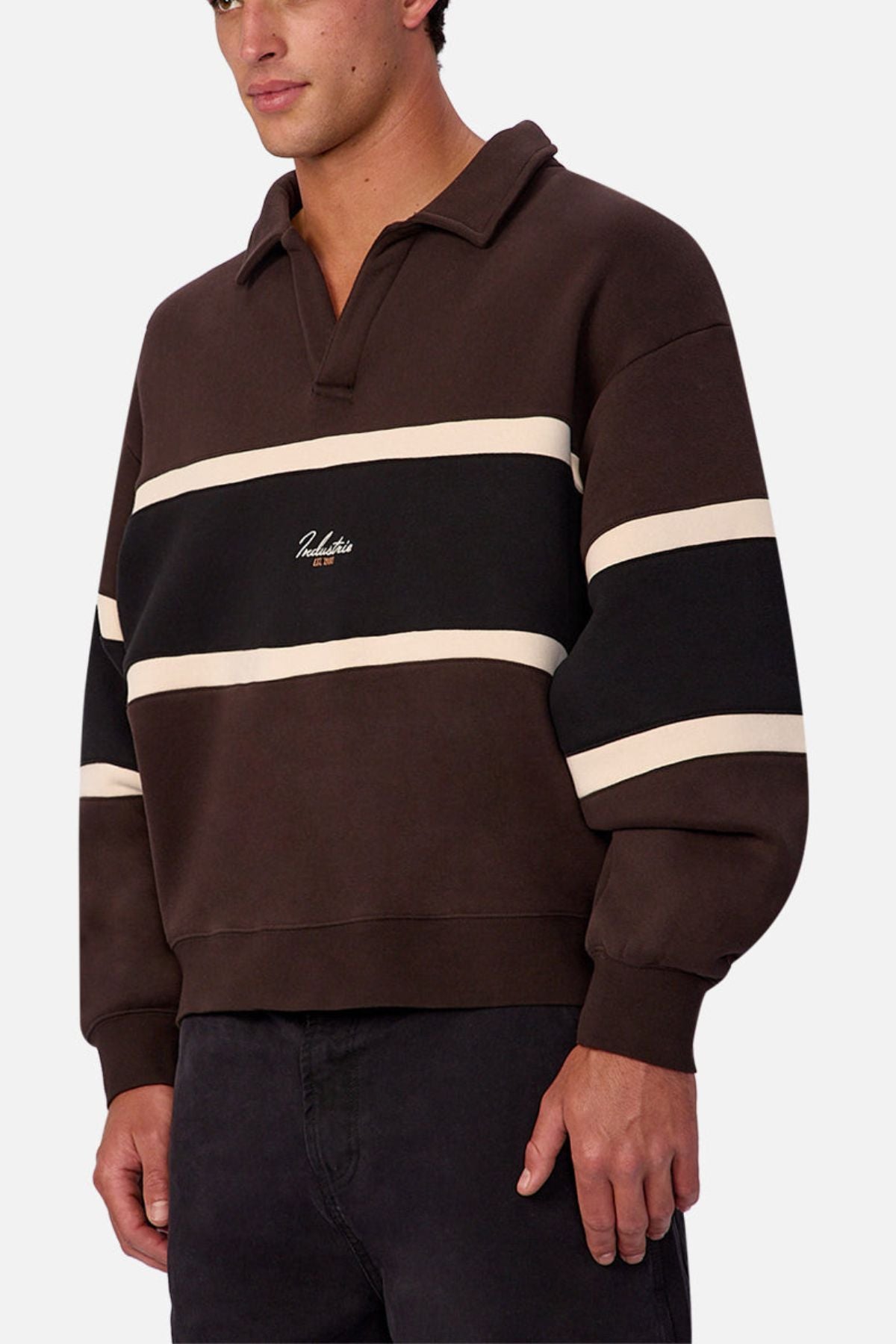 Someone wears the Industrie The Eastbourne Tracktop in dark brown with black and cream horizontal stripes, a camp collar, long sleeves, a chest logo, and a relaxed fit, paired with dark pants.